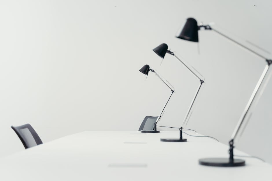 An image of a modern office conference room featuring a long white table with a smooth surface, accompanied by black mesh-back chairs positioned around it. Three sleek, adjustable black desk lamps with chrome arms are evenly spaced along the length of the table, providing directed lighting. The background consists of a plain light-colored wall, free of decorations, creating a minimalistic and tidy atmosphere. The surfaces appear clean and polished, highlighting the importance of surface cleaning and maintenance in a professional setting. The lighting is soft and evenly distributed, emphasizing the cleanliness and order of the room, which reflects services offered by Pimlico Carpet Cleaners in office sanitisation and deep cleaning.