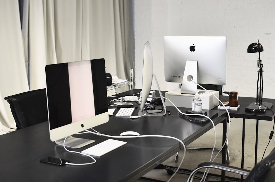 Indoor office workspace with a black desk surface featuring two Apple iMac computers, one with a dark screen and the other with a white screen, along with a wireless keyboard and mouse for each. The desk also holds small office accessories such as a smartphone, stacks of paper, and a bottle of hand sanitizer. In the background, a white brick wall and cream-colored curtains are visible, while a small side table contains a black adjustable desk lamp. The scene is well-lit, clean, and organized, highlighting the surface cleaning and maintenance of a modern office environment, as managed by Pimlico Carpet Cleaners from the page titled 'Office Cleaning Options by Tate Britain, Pimlico, PIMLICO' at pimlicocarpetcleaners.co.uk.