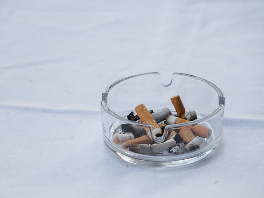 A close-up of a person's hand holding a lit cigarette, with smoke rising upwards in a residential setting. The background features a beige wooden door or panel, with a smooth, clean surface and soft i